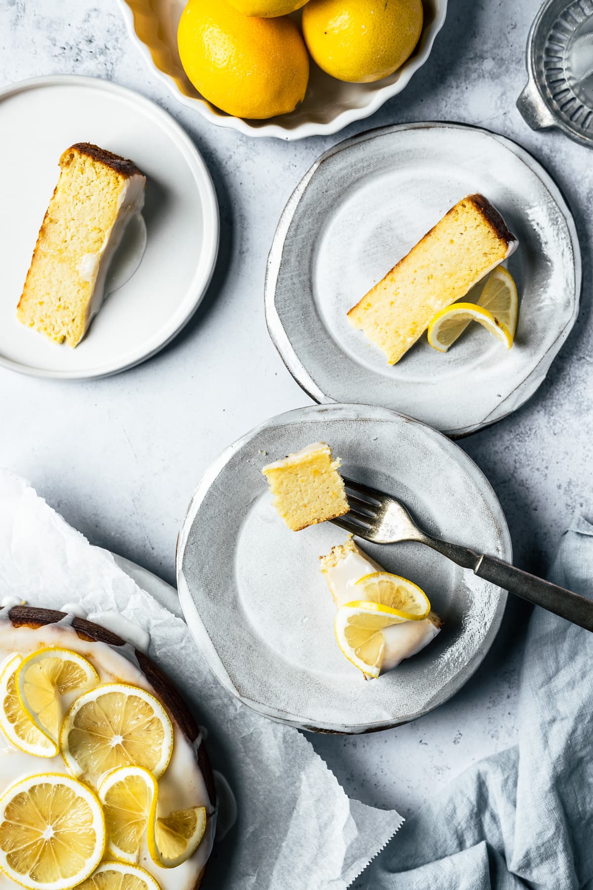 Three white ceramic plates hold slices of yellow cake with white glaze and lemon twists on a light blue concrete surface. There is a bite of cake on a fork on one of the plates. The rest of the lemon decorated cake peeks into the frame at bottom left, and a bowl of lemons and a juicer are partially in the frame at top.