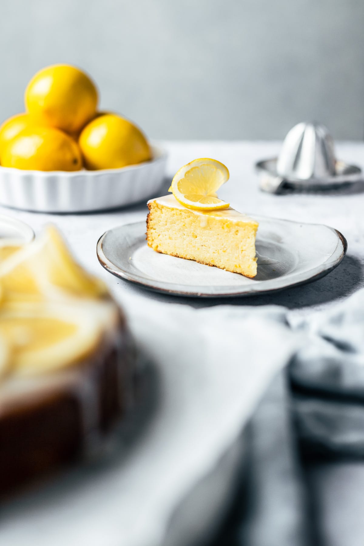 A front view of a slice of cake with a twisted lemon slice decoration on top, on a white ceramic plate. Lemons, a juicer, the rest of the cake, and a light blue napkin all surround the cake, slightly out of focus.