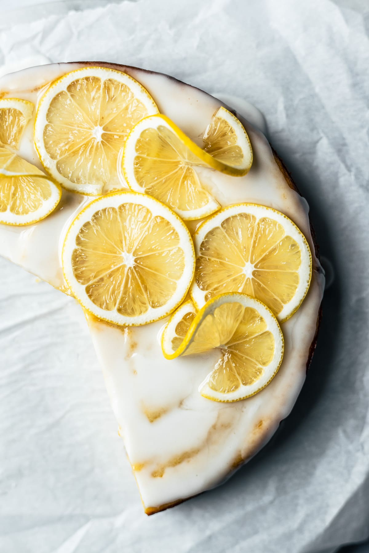 Top view of glazed cake with lemon slices on top. There are slices missing from the cake, and it rests on a white parchment surface.