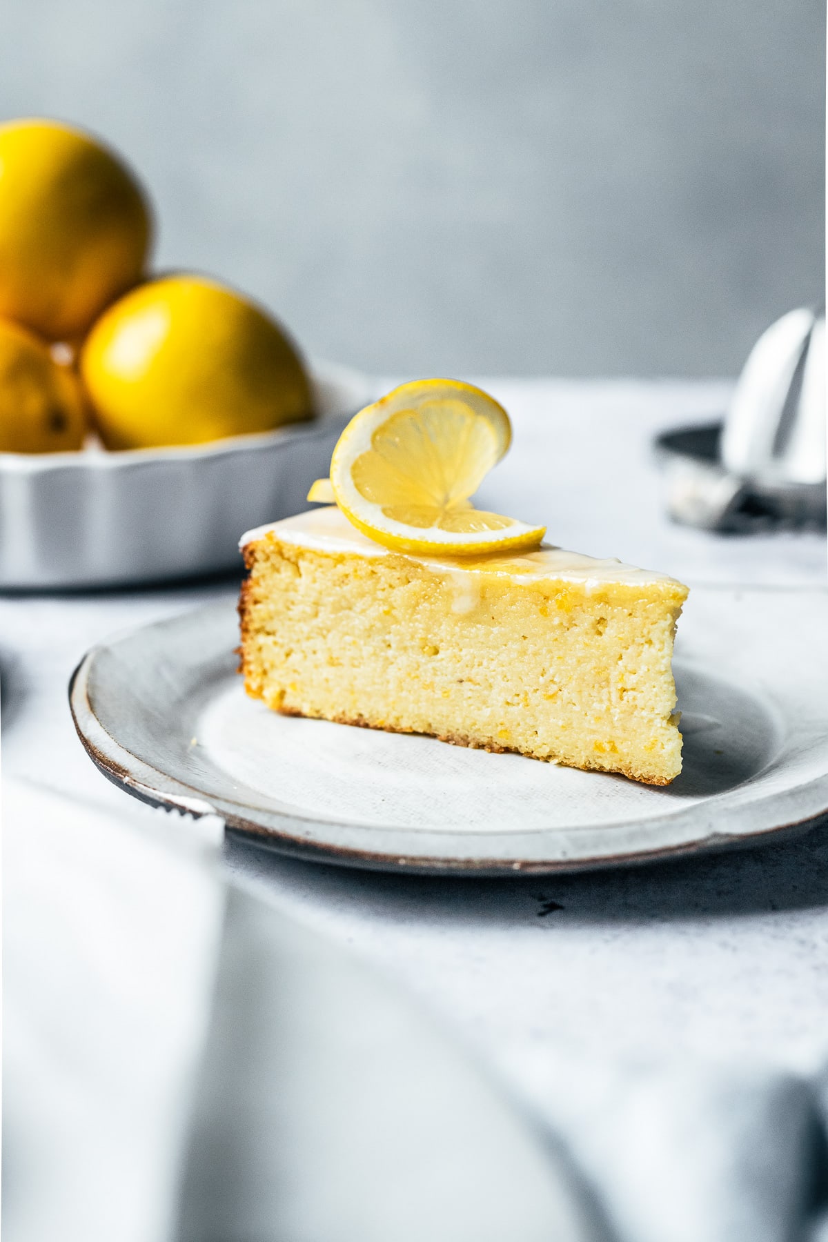 A close up side view of a slice of cake with a twisted lemon slice decoration on top, on a white ceramic plate. Lemons, a juicer, and a light blue napkin all surround the cake, slightly out of focus.