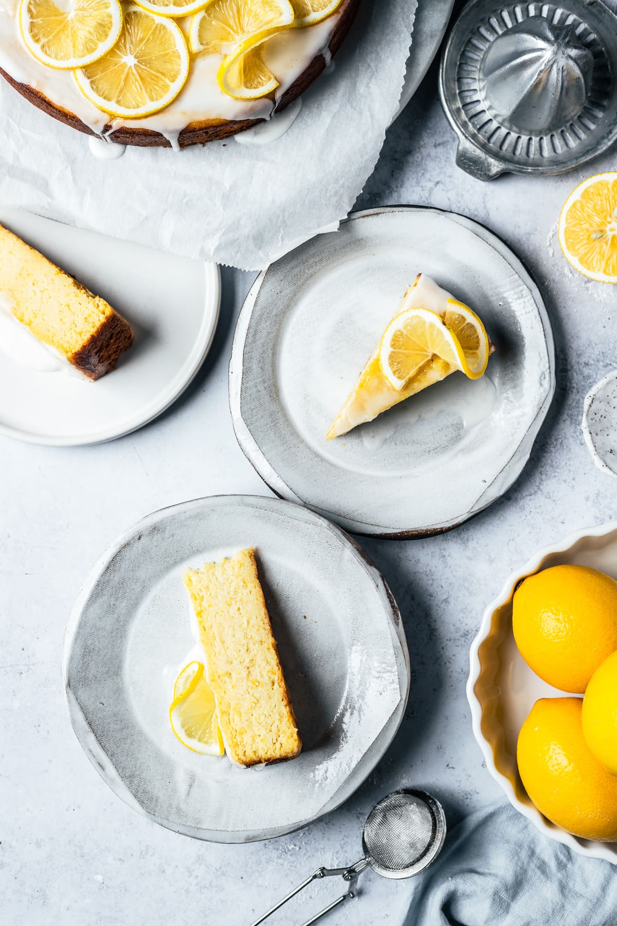 Three white ceramic plates hold slices of yellow cake with white glaze and lemon twists on a light blue concrete surface. A white bowl of lemons peeks into the frame at bottom right, and the rest of the cake and a juicer are partially in the frame at top.