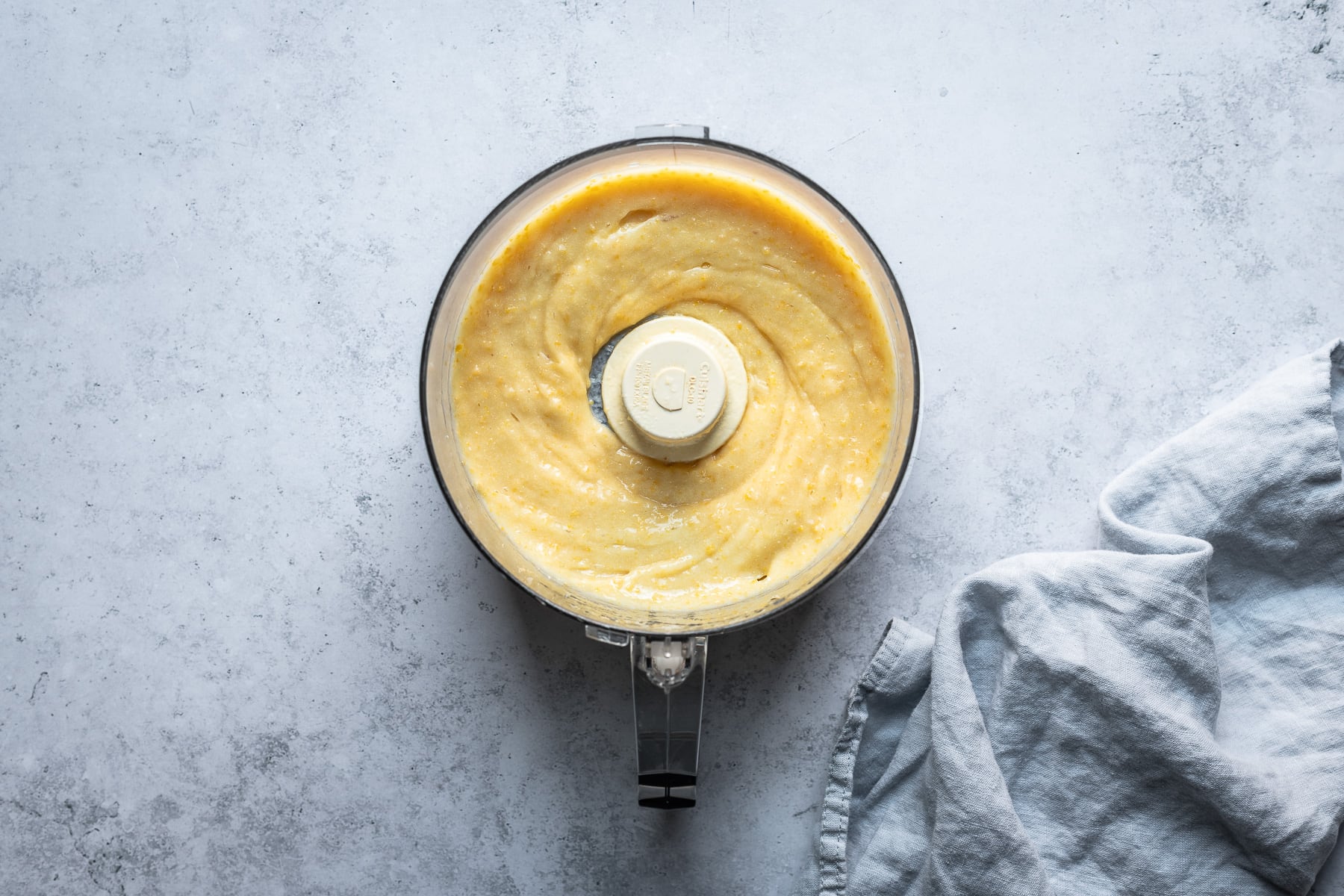 The bowl of a food processor with a bright lemon puree inside. There is a light blue concrete surface in the background with a light blue napkin at right.