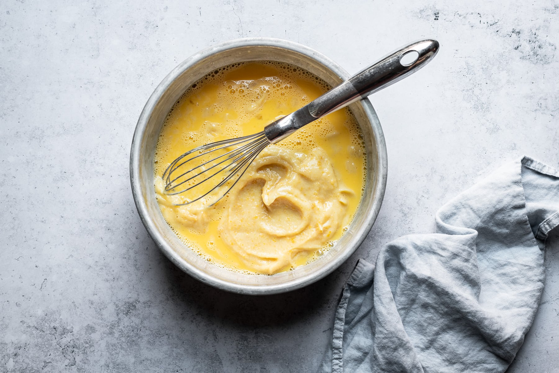 A process shot showing a bowl of eggs and lemon with a whisk resting in it. The bowl rests on a light blue concrete background with a light blue napkin resting at right.