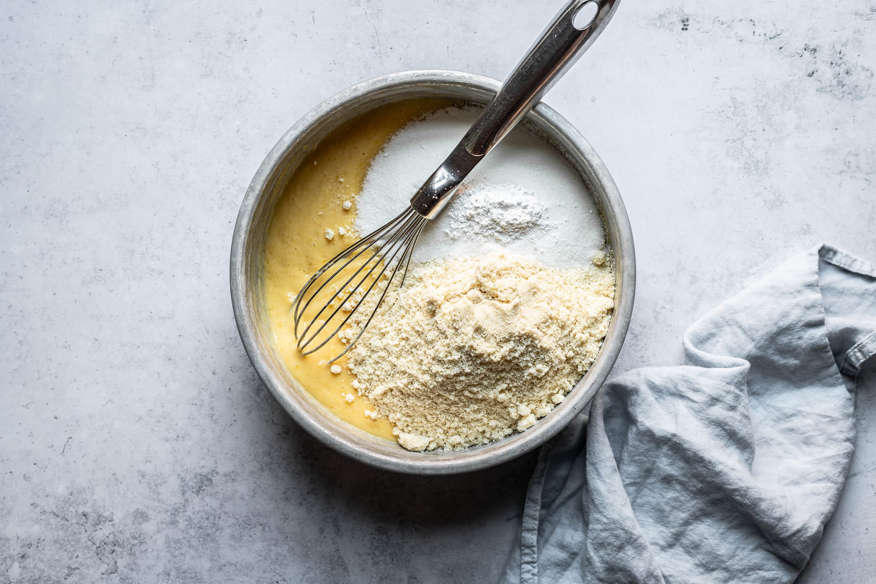 A process shot showing a bowl of eggs, lemon, almond flour and sugar with a whisk resting in it. The bowl rests on a light blue concrete background with a light blue napkin at right.