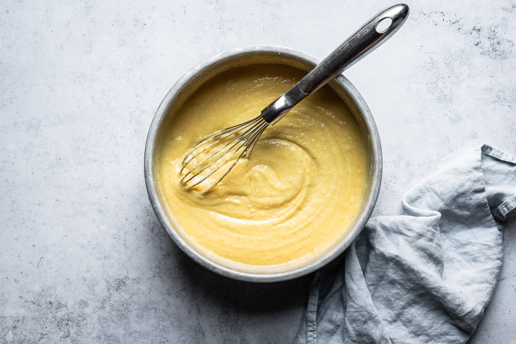 A process shot showing a bowl filled with pale yellow cake batter and a whisk. The pan rests on a light blue concrete background with a light blue napkin resting at right.