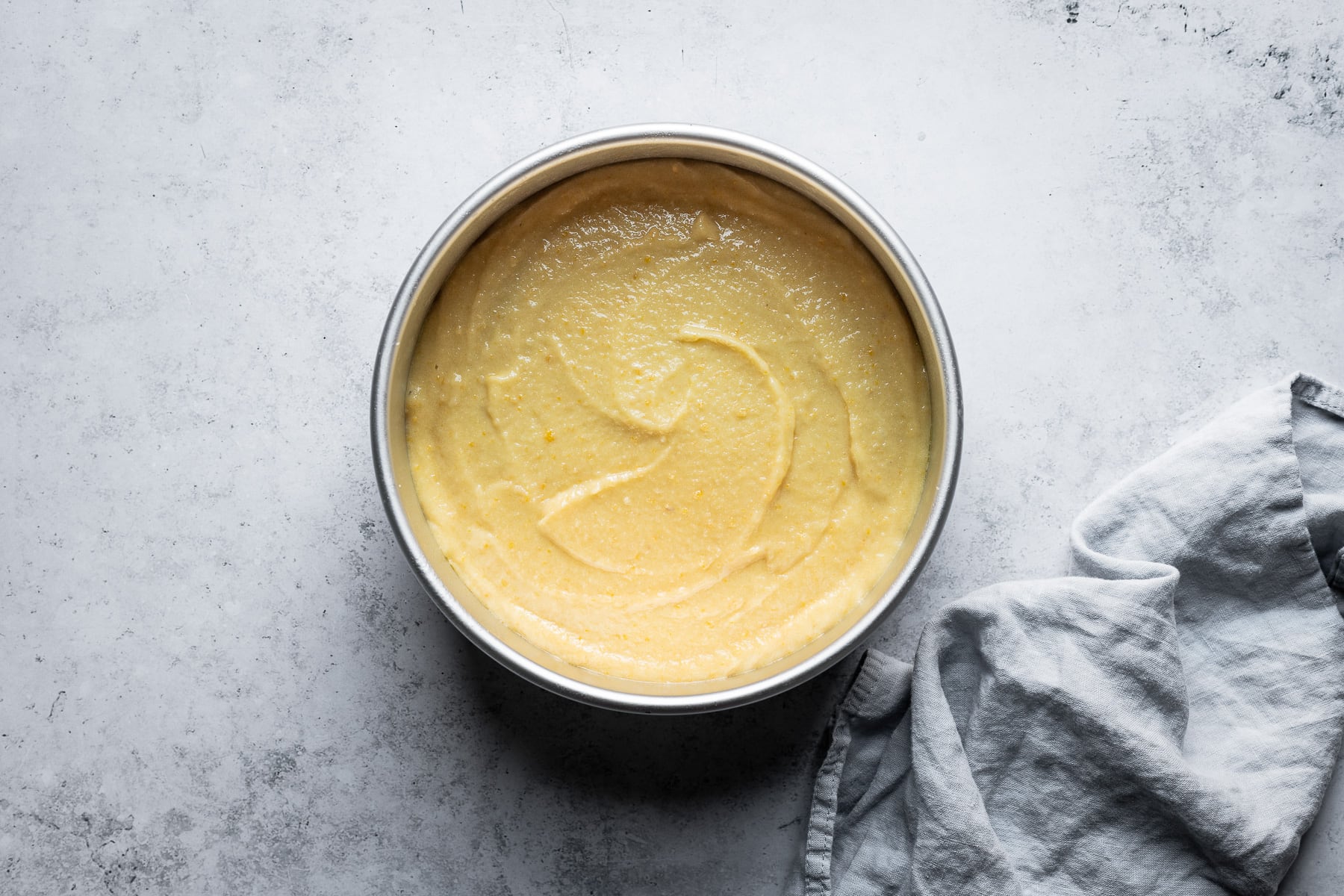 A process shot showing a cake pan filled with pale yellow batter. The pan rests on a light blue concrete background with a light blue napkin resting at right.