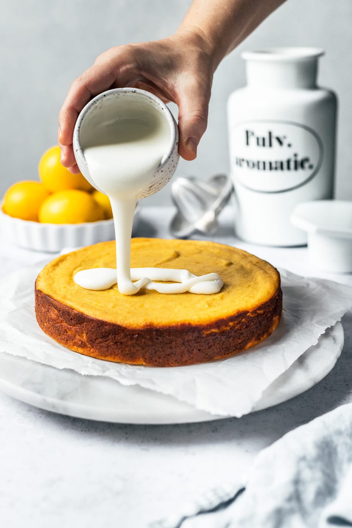 A hand pours a white glaze from a white speckled ceramic container onto a vibrant yellow cake, which rests on parchment paper and a marble plate. There are bright yellow lemons, a juicer, and a porcelain apothecary container in the background.