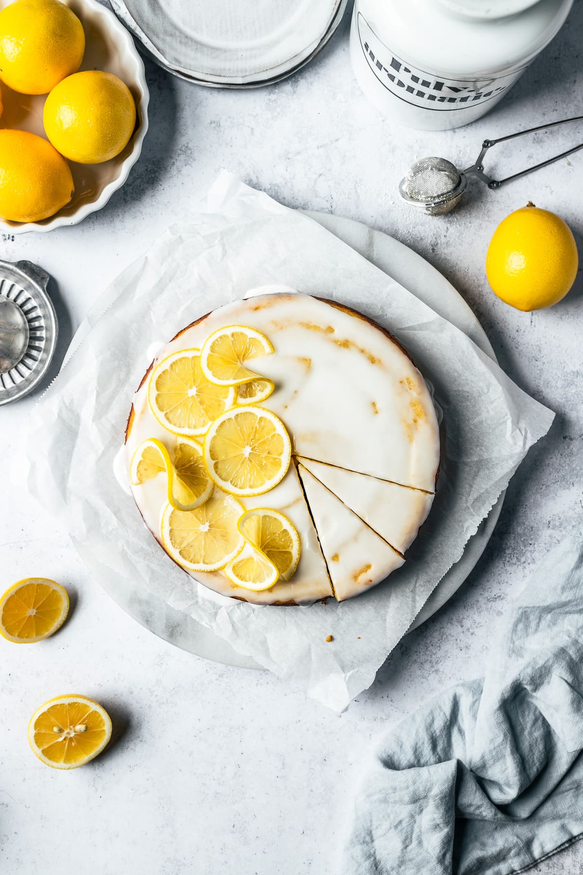 A yellow cake with white glaze, decorated with thin lemon slices. There are two slices marked in the cake. The cake rests on a white marble platter on white parchment paper on a light blue concrete background. Lemons, a juicer, and a light blue napkin peek in from the edges of the photo.