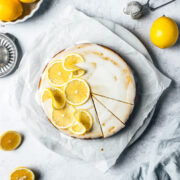 A yellow cake with white glaze, decorated with thin lemon slices. There are two slices marked in the cake. The cake rests on a white marble platter on white parchment paper on a light blue concrete background. Lemons, a juicer, and a light blue napkin peek in from the edges of the photo.