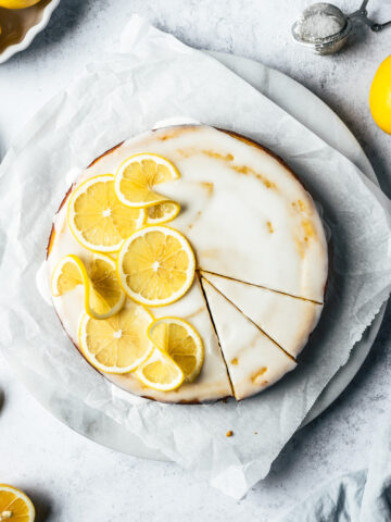 A yellow cake with white glaze, decorated with thin lemon slices. There are two slices marked in the cake. The cake rests on a white marble platter on white parchment paper on a light blue concrete background. Lemons, a juicer, and a light blue napkin peek in from the edges of the photo.