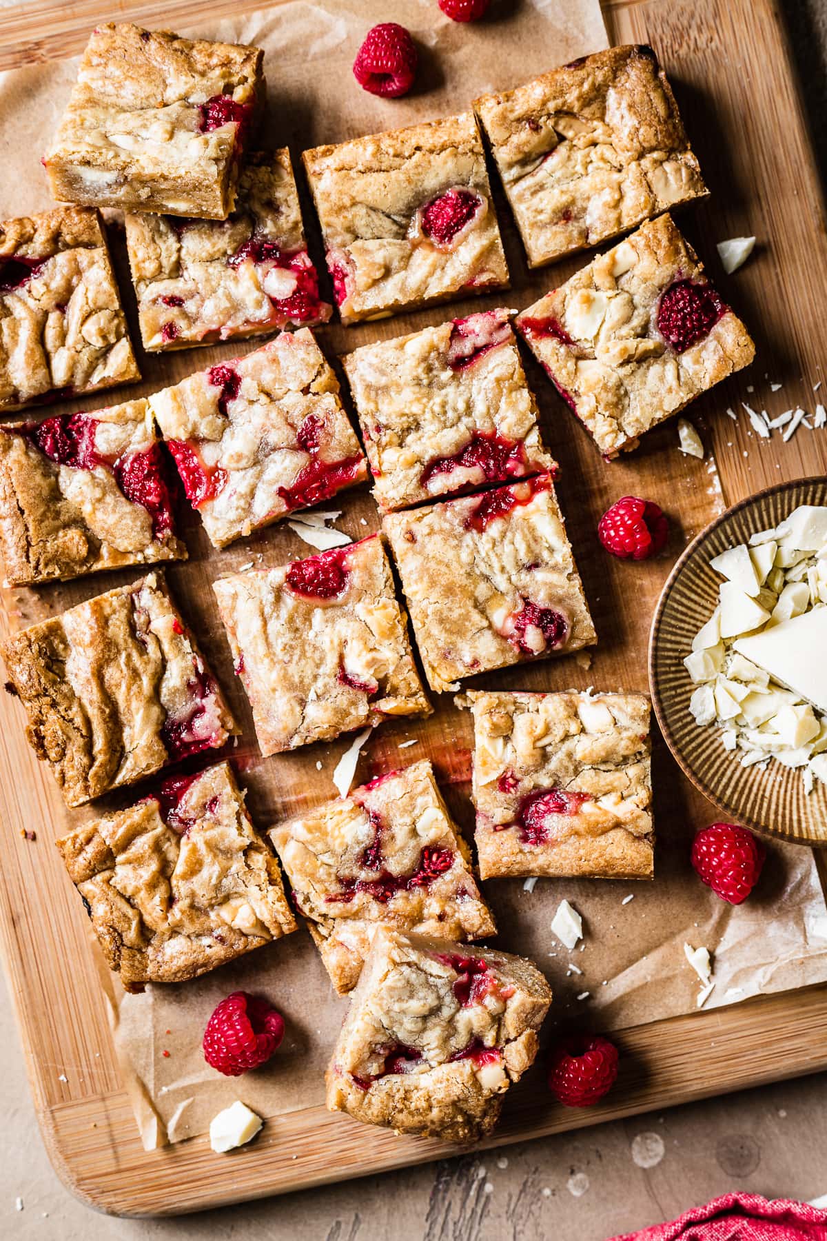 Raspberry and white chocolate stuffed bar cookies cut into squares on a wooden cutting board. Raspberries and white chocolate peek in from the edge of the image.
