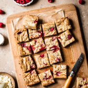 Raspberry stuffed bars cut into squares on a wooden cutting board with knife resting nearby. Small bowls of raspberries and white chocolate peek in from the edge of the image.