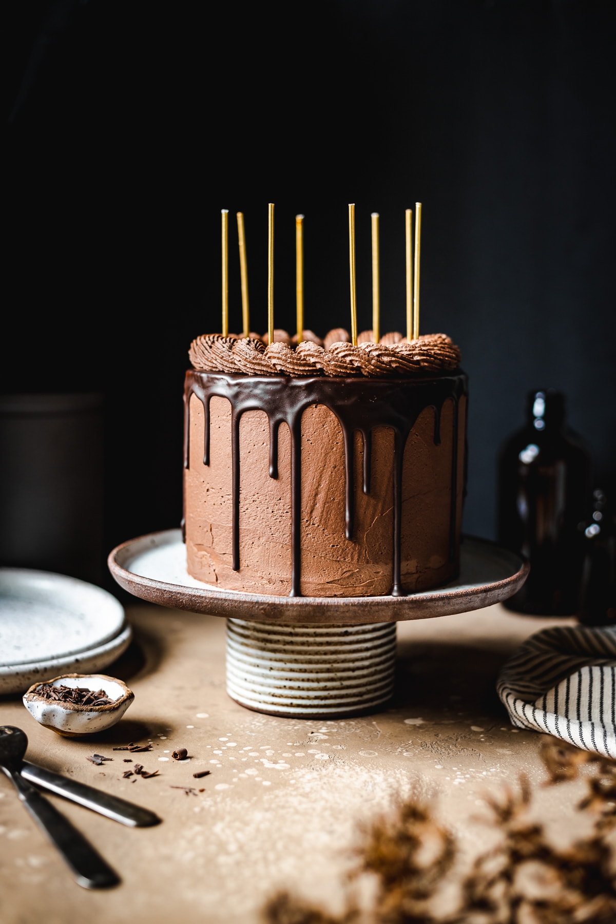 A moody image of a drip cake on a ceramic cake stand on a tan stone surface. There are yellow candles on the cake. Plates, a napkin, and forks rest nearby. The background is black.