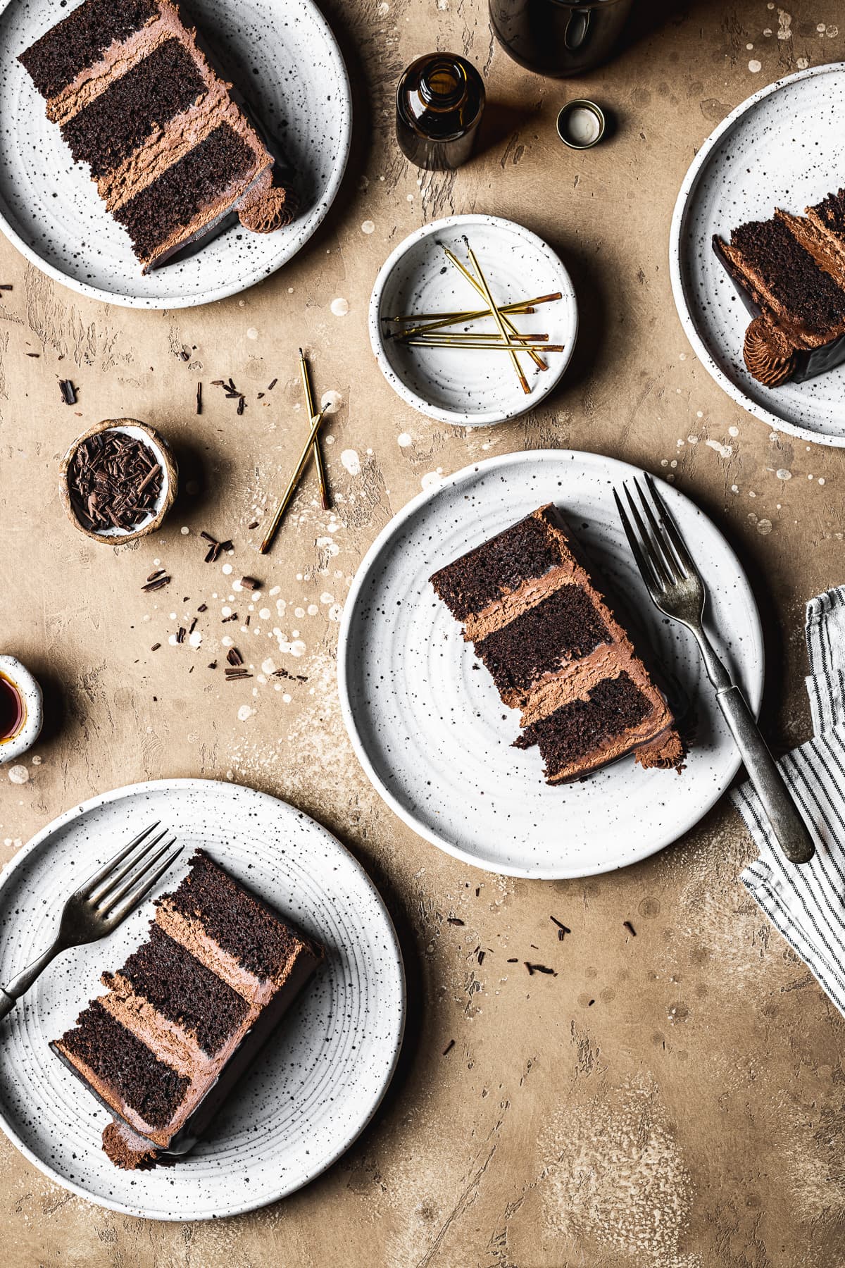 Four slices of chocolate cake on white ceramic plates on a tan stone surface. There is a bowl of candles nearby, along with amber glass bottles, a striped napkin and small bowls.