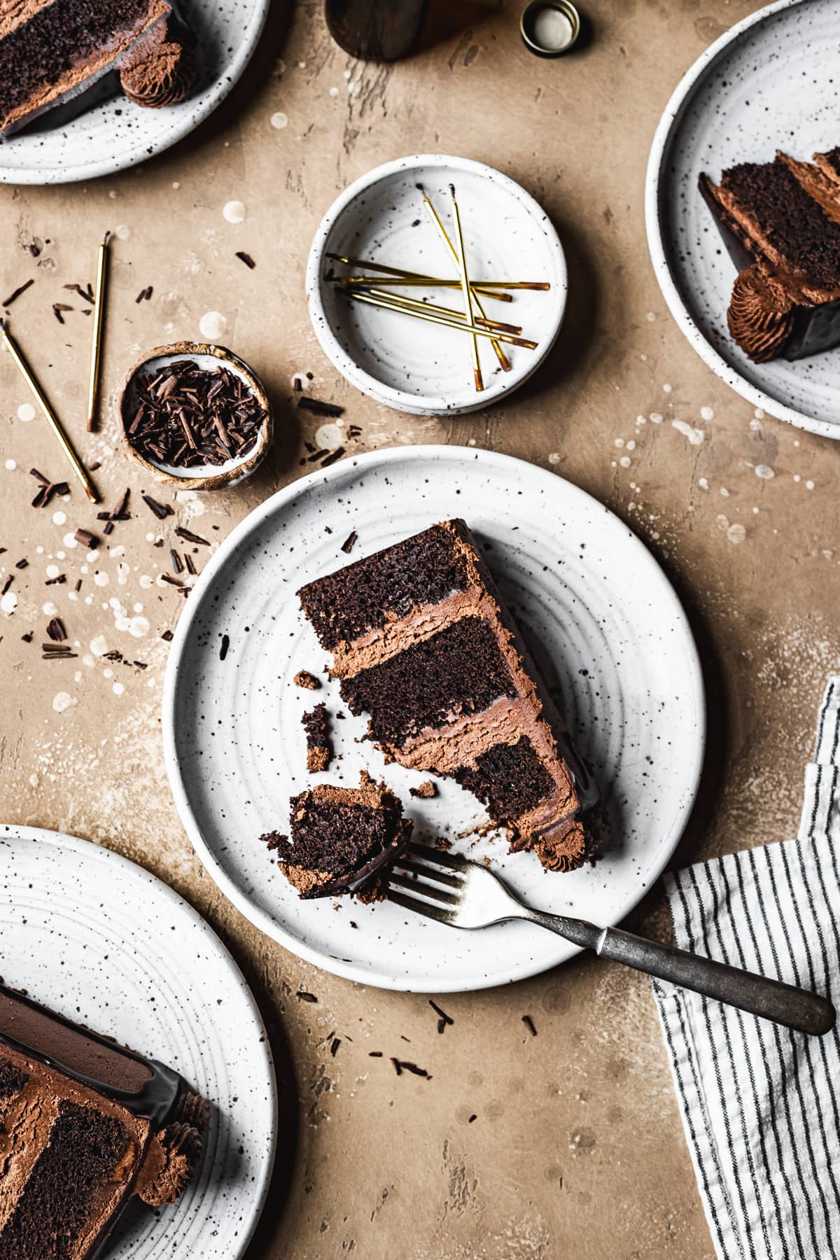 A slice of chocolate cake on a white ceramic plate on a tan stone surface. There is a bowl of candles nearby, a striped napkin and small bowls filled with candles and chocolate shavings.