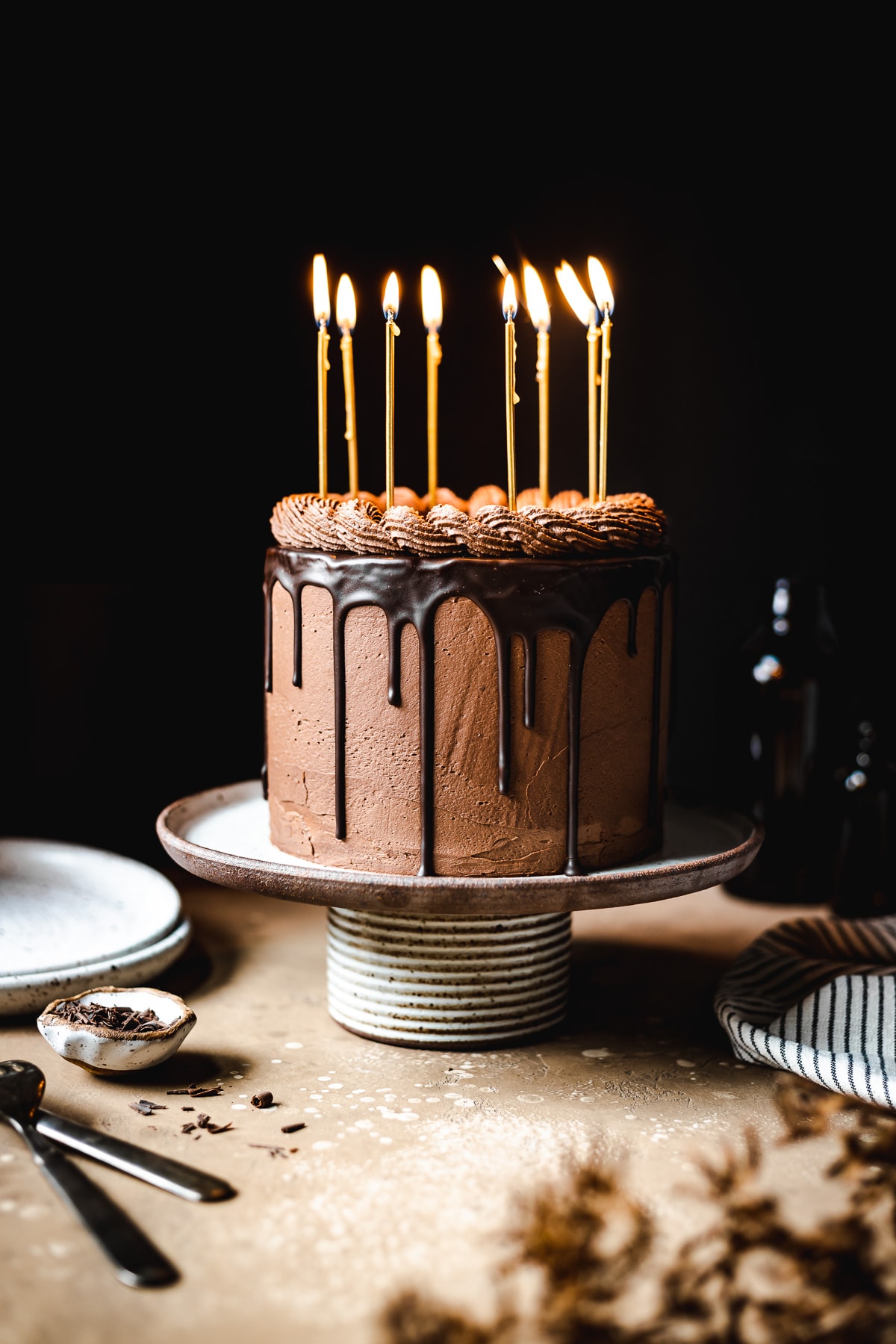 A moody image of a drip cake on a ceramic cake stand on a tan stone surface. The candles on the cake are lit. Plates, a napkin, and forks rest nearby. The background is black.