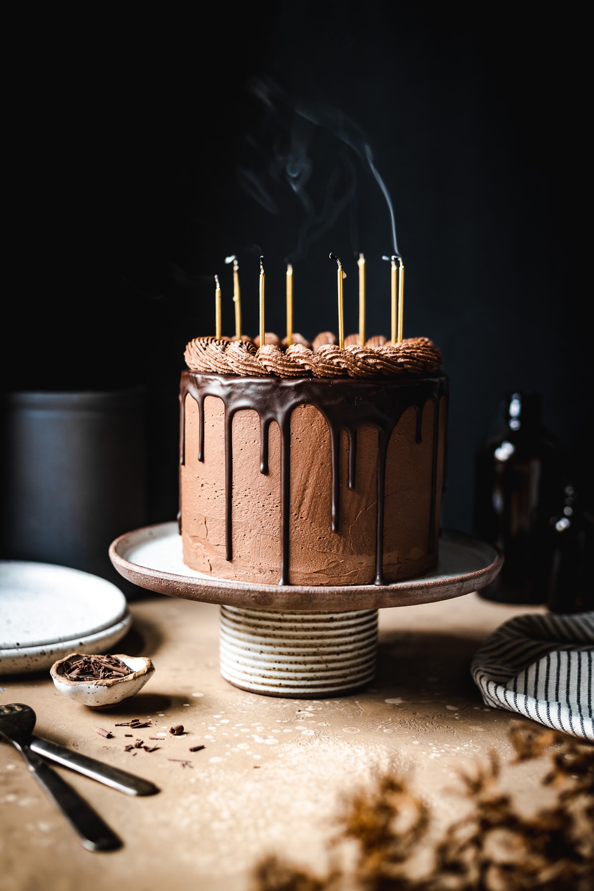 A moody image of a drip cake on a ceramic cake stand on a tan stone surface. Smoke curls up from candles on the cake that were just blown out. Plates, a napkin, and forks rest nearby. The background is black.