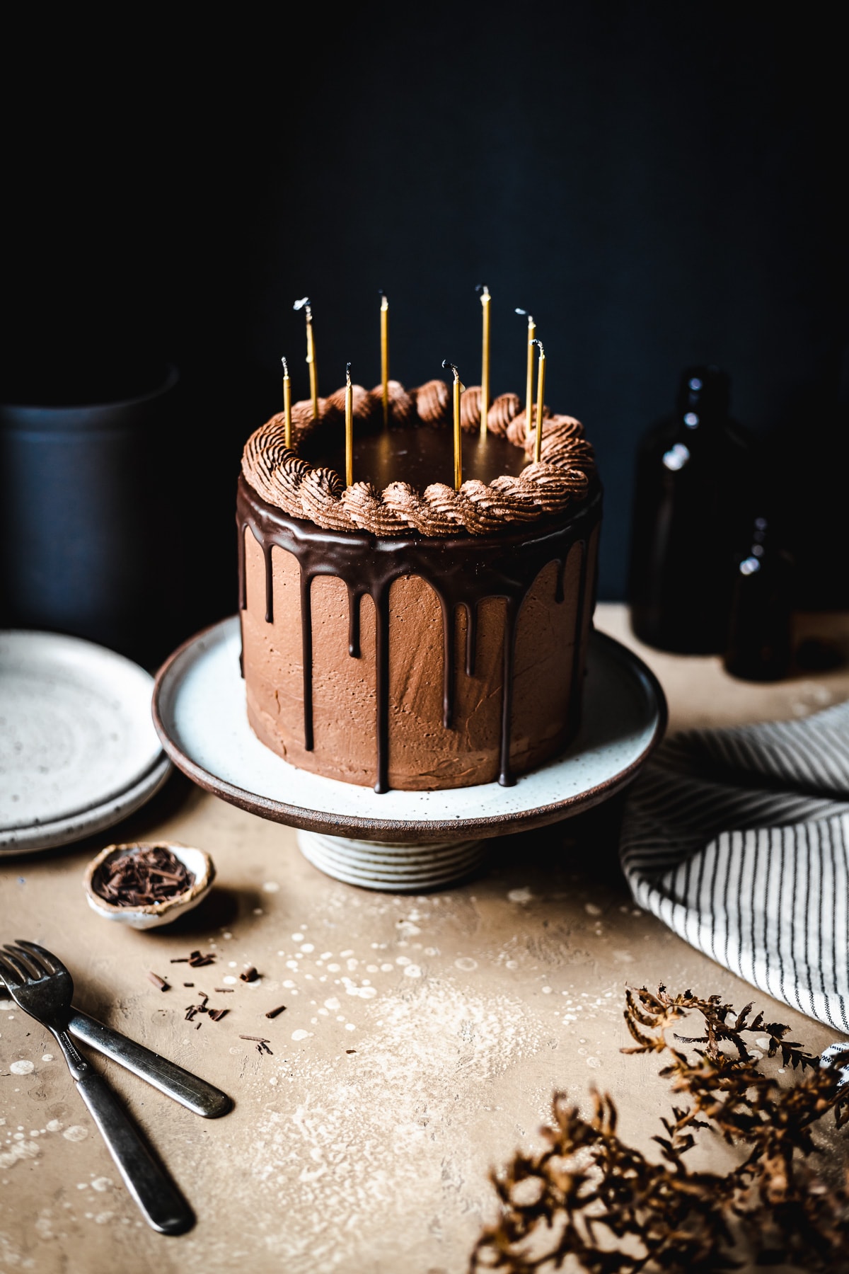 A moody image of a drip cake on a ceramic cake stand on a tan stone surface. There are candles on the cake. Plates, a napkin, and forks rest nearby. The background is black.