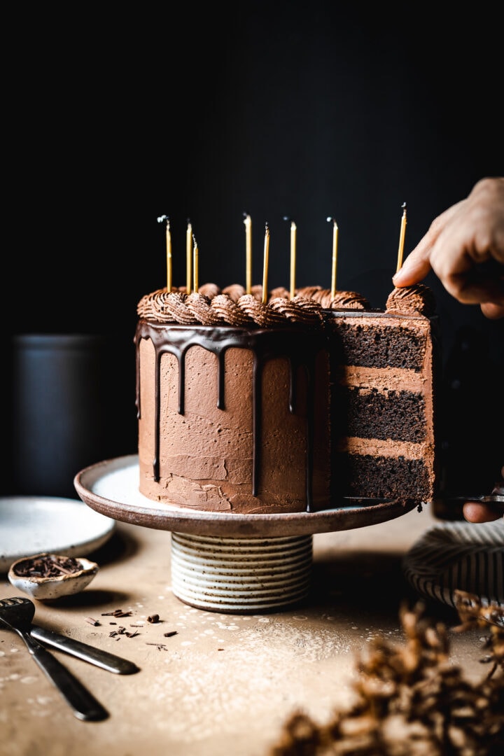 A moody image of hands removing a slice of cake from a ceramic cake stand on a tan stone surface. There are candles on the cake. Plates, a napkin, and forks rest nearby. The background is black.