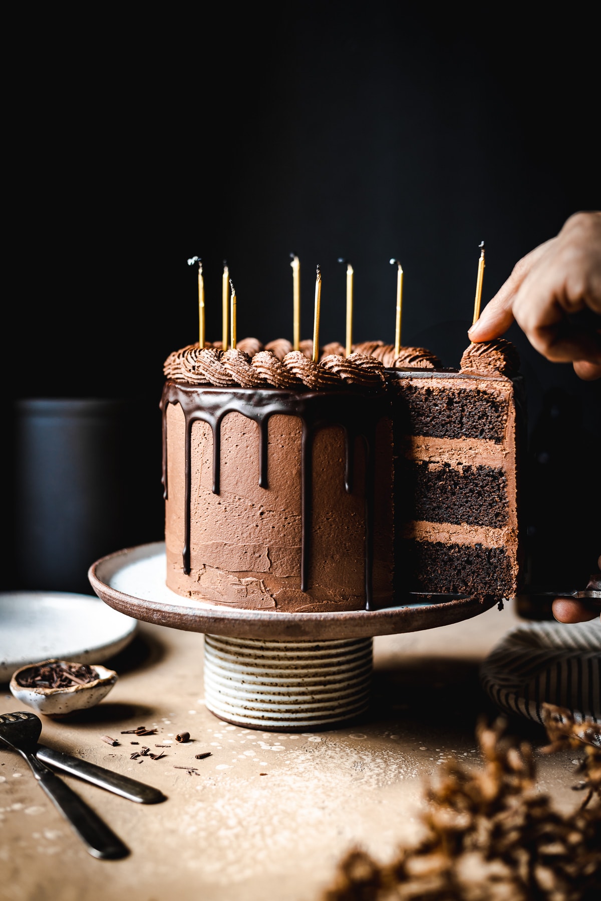 A moody image of hands removing a slice of chocolate cake from a ceramic cake stand on a tan stone surface. There are candles on the cake. Plates, a napkin, and forks rest nearby. The background is black.