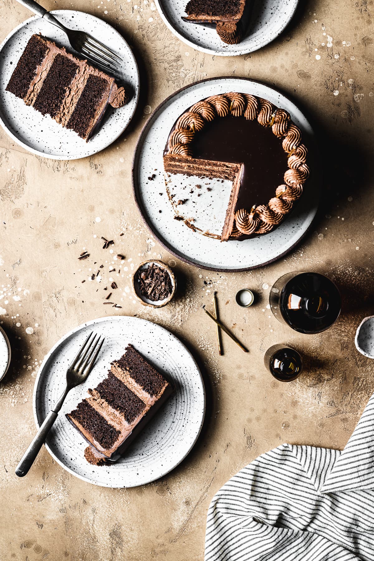 A triple chocolate cake on a white ceramic plate is cut into slices on surrounding plates on a tan stone surface. There are candles, amber glass jars, a striped napkin and small bowls nearby.