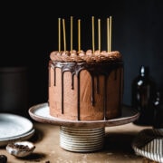 A moody image of a cake on a ceramic cake stand on a tan stone surface. There are yellow candles on the cake. Plates, a napkin, and forks rest nearby. The background is black.