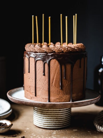 A moody image of a cake on a ceramic cake stand on a tan stone surface. There are yellow candles on the cake. Plates, a napkin, and forks rest nearby. The background is black.