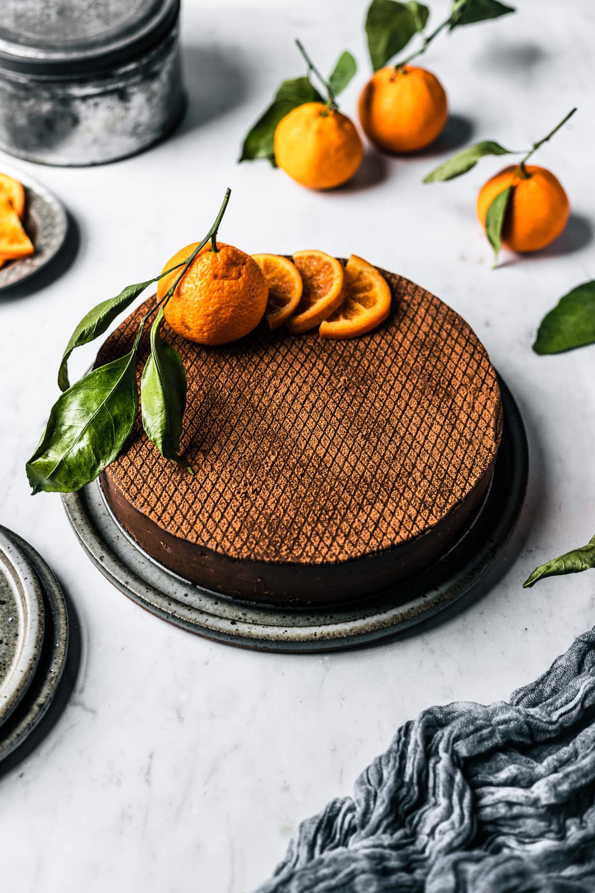 An angled view of a cheesecake decorated with orange slices and a whole orange with a few slices removed, on a white marble surface. There are more oranges with leaves nearby, a metal tin, and a plate of sliced candied oranges.