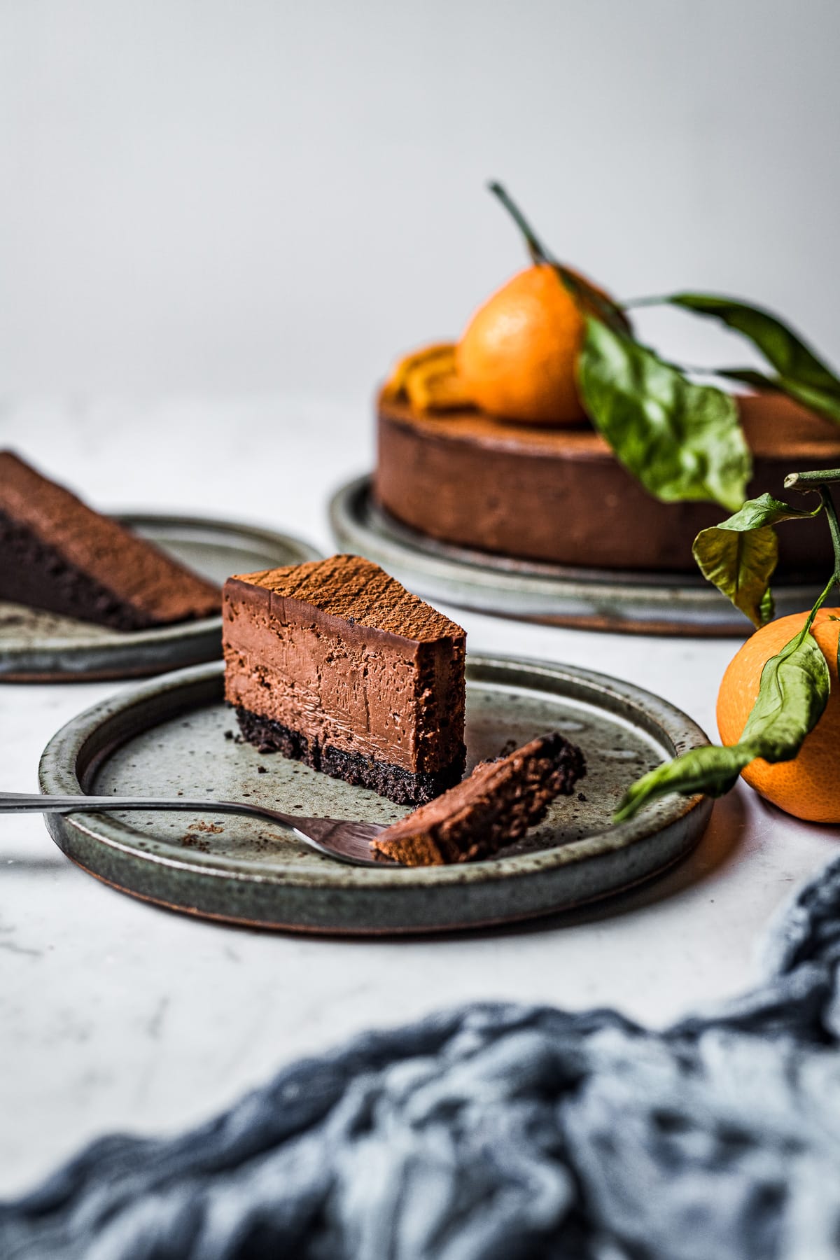 A slice of cheesecake on a ceramic plate with a bite resting on a fork. The rest of the cheesecake is in the background at right, and there are mandarin oranges with leaves resting nearby, along with a grey blue linen.