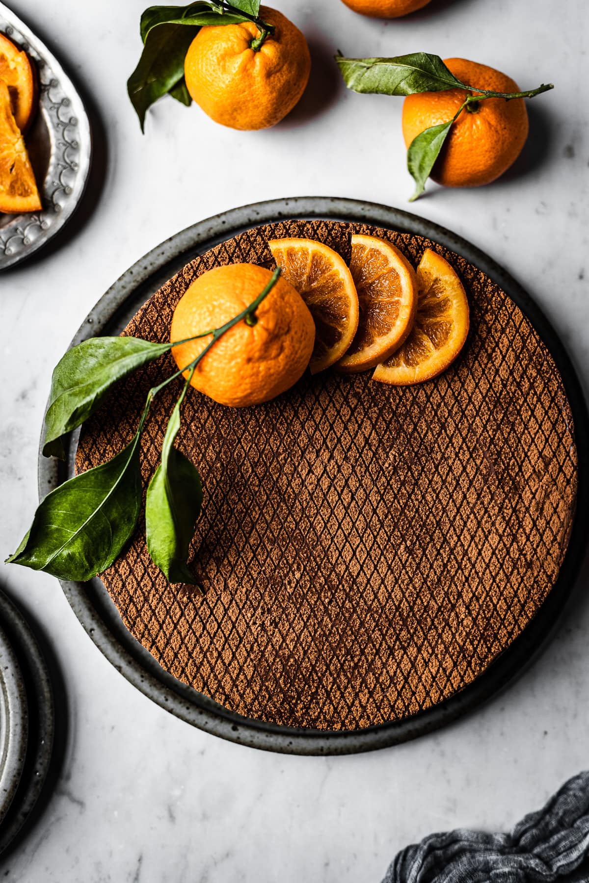 A chocolate orange cheesecake decorated with a stenciled grid of cocoa powder, orange slices and a whole orange on a white marble surface. There are more oranges with leaves nearby, and a plate of sliced candied oranges.