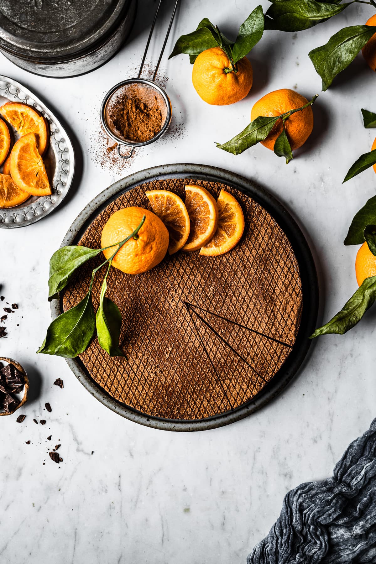 A chocolate orange cheesecake decorated with orange slices and a whole orange with a few slices removed, on a white marble surface. There are more oranges with leaves nearby, a small bowl of chopped chocolate, and a plate of sliced candied oranges.