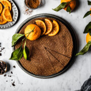 A chocolate orange cheesecake decorated with orange slices and a whole orange with a few slices removed, on a white marble surface. There are more oranges with leaves nearby, a small bowl of chopped chocolate, and a plate of sliced candied oranges.