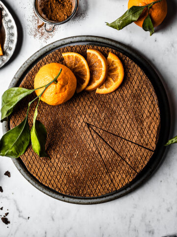 A chocolate orange cheesecake decorated with orange slices and a whole orange with a few slices removed, on a white marble surface. There are more oranges with leaves nearby, a small bowl of chopped chocolate, and a plate of sliced candied oranges.