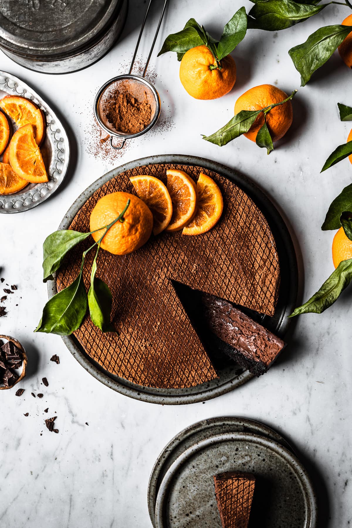 A chocolate orange cheesecake decorated with orange slices and a whole orange with a few slices removed, on a white marble surface. There are more oranges with leaves nearby, a small bowl of chopped chocolate, a metal tin, and a plate of sliced candied oranges.