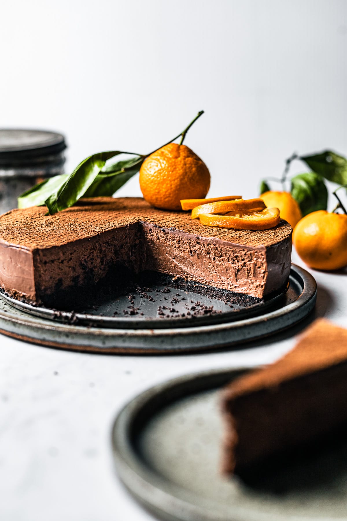 A chocolate orange cheesecake that has had slices removed, revealing the creamy interior and cocoa crust. It is decorated with orange slices and a whole orange. A slice of cheesecake is out of focus at front right and there are more oranges in the background at right.