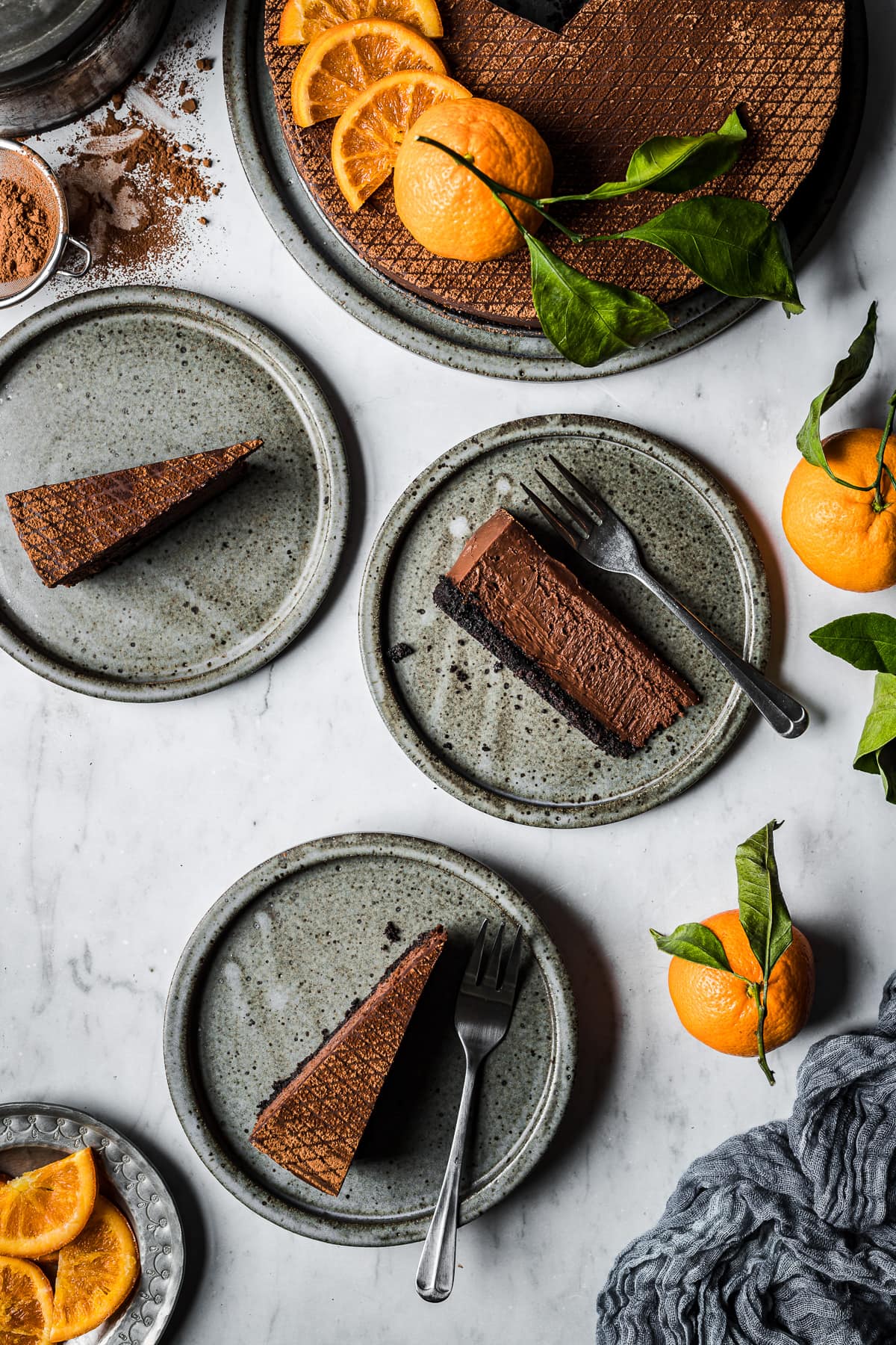 Slices of cheesecake on grey ceramic plates on a marble surface. The larger cheesecake is on a platter at top right. There are oranges, a blue linen, and a small strainer of cocoa powder nearby.