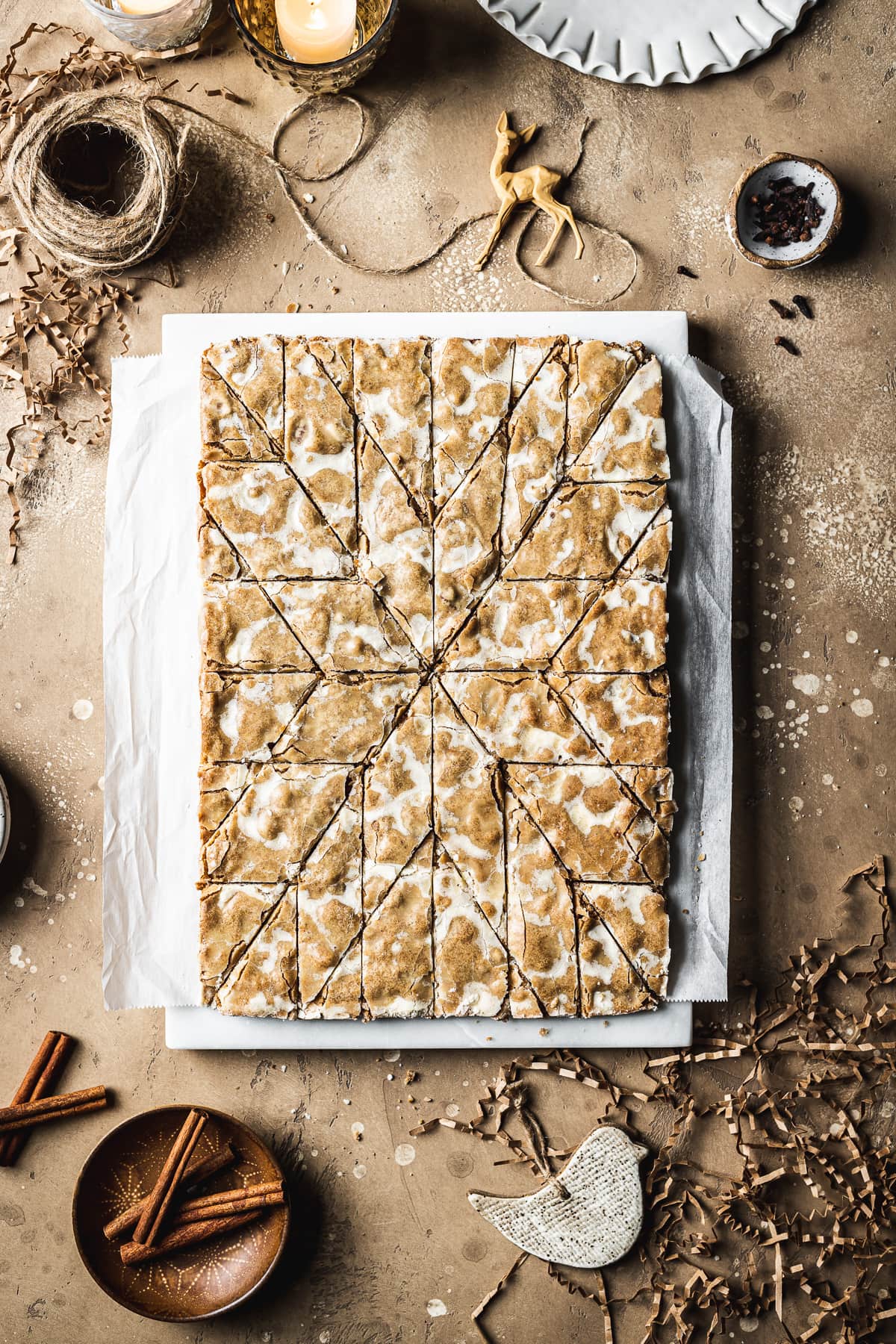 German cookies on a marble block and white parchment paper, cut into a decorative holiday star pattern. The marble block rests on a tan stone surface and is surrounded by cinnamon sticks, cloves, a votive candle, brown twine, holiday figurines and more cookies.