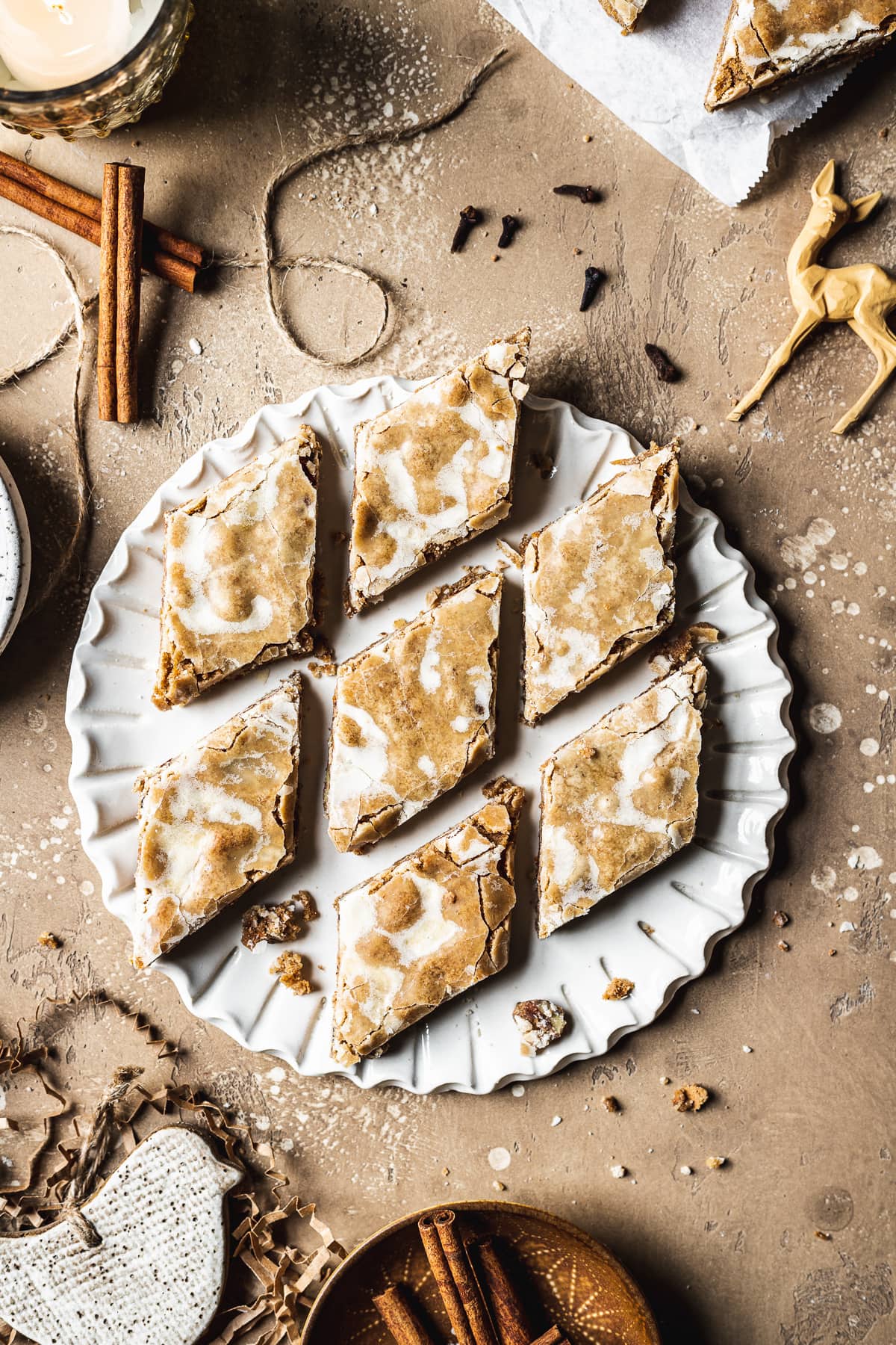 A fluted white ceramic plate holds diamond shaped glazed bar cookies. The plate is on a tan stone surface and is surrounded by cinnamon sticks, cloves, a votive candle, holiday figurines and more cookies.