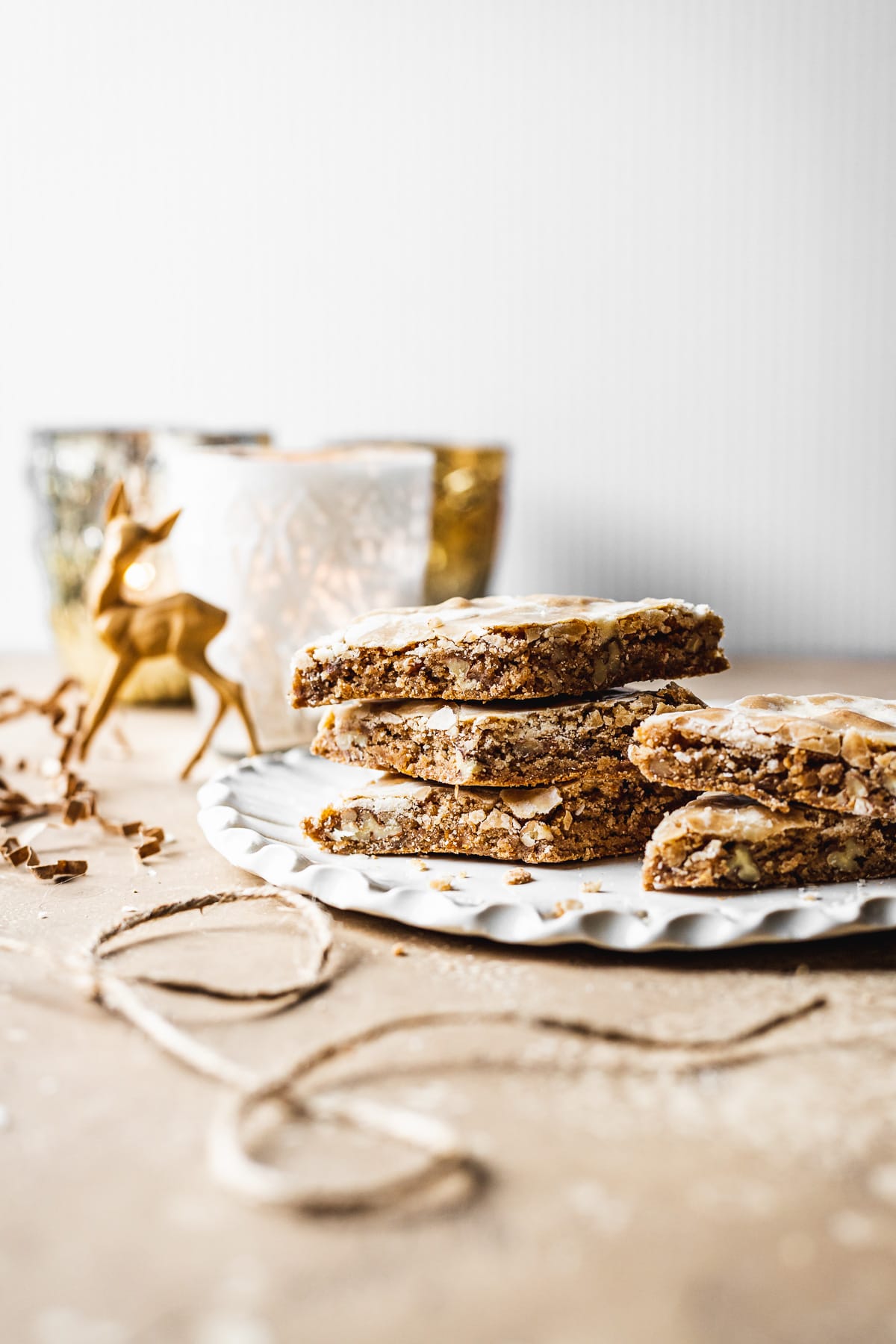 A stack of bar cookies on a fluted white ceramic plate on a tan surface. Brown twine, a reindeer figurine and mercury glass votives surround the plate.