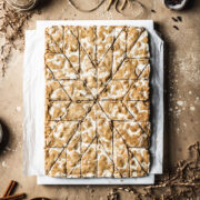 German bar cookies on a marble block and white parchment paper, cut into a decorative holiday star pattern. The marble block rests on a tan stone surface and is surrounded by cinnamon sticks, cloves, a votive candle, brown twine, holiday figurines and more cookies.