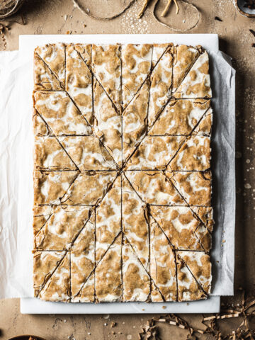 German bar cookies on a marble block and white parchment paper, cut into a decorative holiday star pattern. The marble block rests on a tan stone surface and is surrounded by cinnamon sticks, cloves, a votive candle, brown twine, holiday figurines and more cookies.