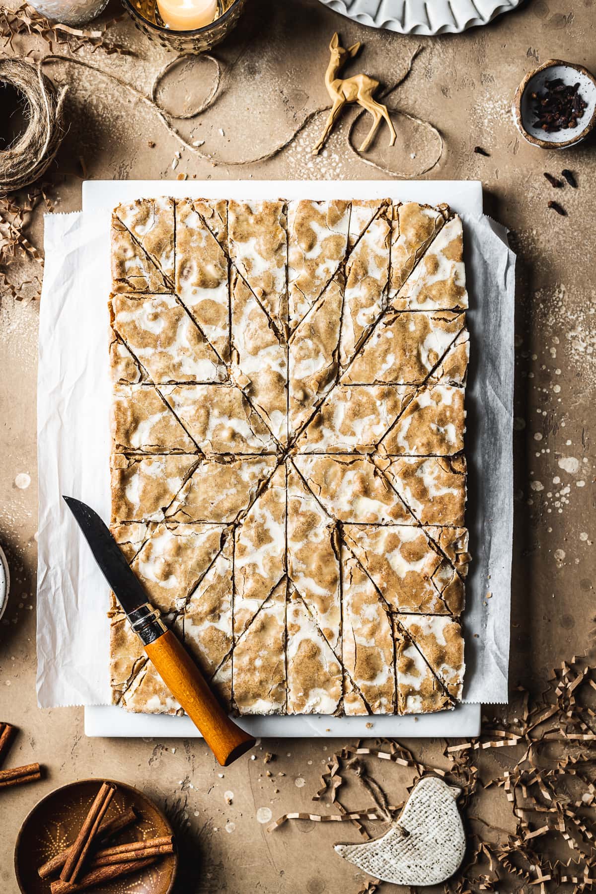 Bar cookies on a marble block and white parchment paper, cut into a decorative holiday star pattern. The marble block rests on a tan stone surface and is surrounded by cinnamon sticks, cloves, a votive candle, brown twine, holiday figurines and more cookies.