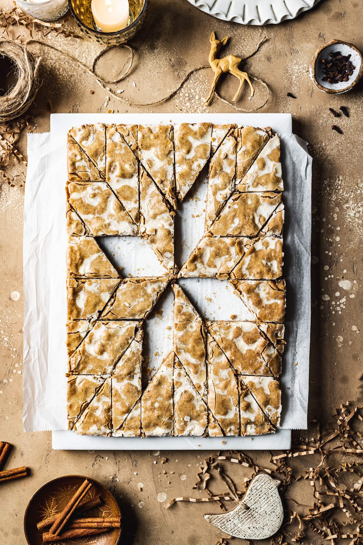 Bar cookies on a marble block and white parchment paper, cut into a decorative holiday star pattern. Four of the cookies in the middle have been removed, revealing the white parchment paper below. The marble block rests on a tan stone surface and is surrounded by cinnamon sticks, cloves, a votive candle, brown twine, holiday figurines and more cookies.