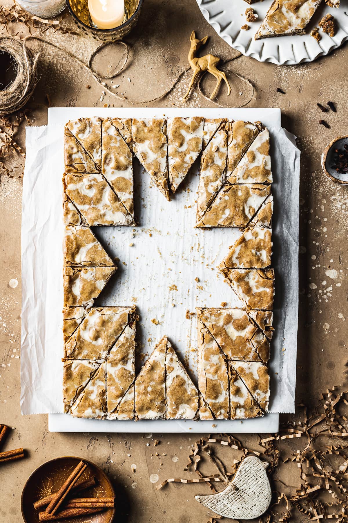 Bar cookies on a marble block and white parchment paper, cut into a decorative holiday star pattern. The cookies in the middle have been removed, revealing the white parchment paper below. The marble block rests on a tan stone surface and is surrounded by cinnamon sticks, cloves, a votive candle, brown twine, holiday figurines and more cookies.