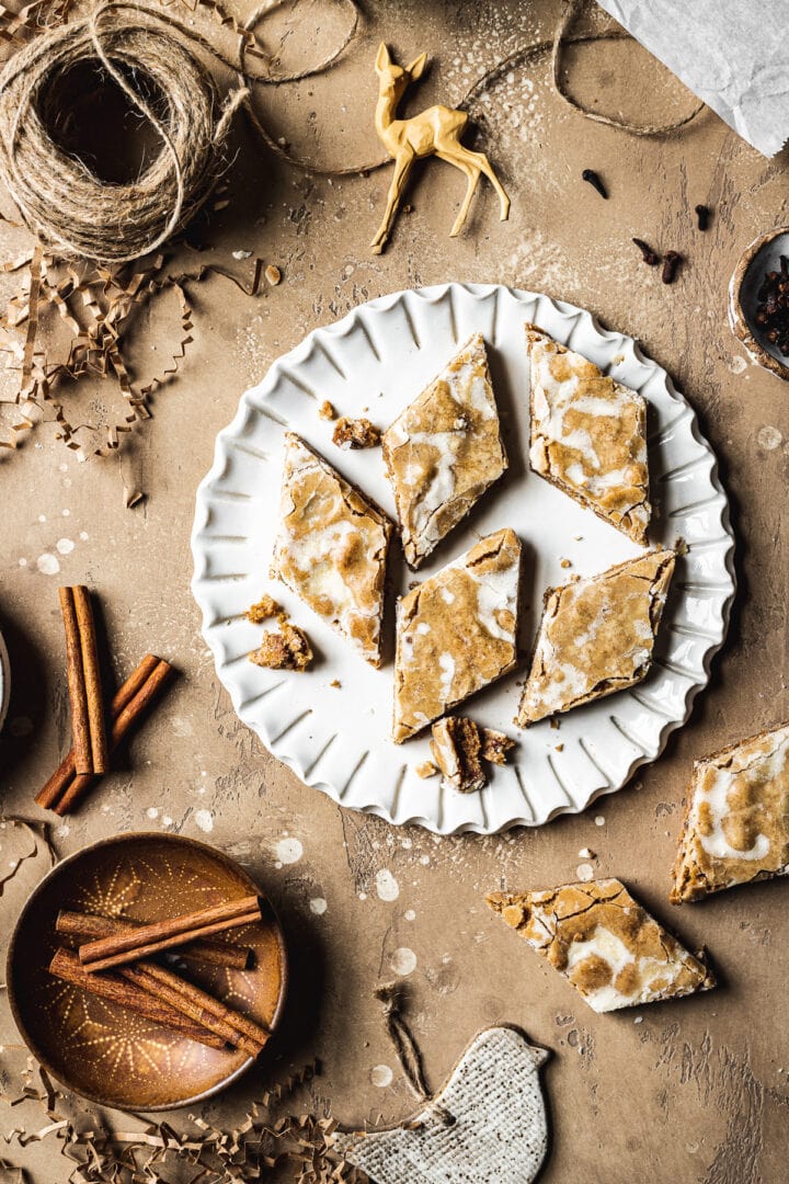 A fluted white ceramic plate holds diamond shaped glazed bar cookies. The plate is on a tan stone surface and is surrounded by cinnamon sticks, cloves, a votive candle, brown twine, holiday figurines and more cookies.