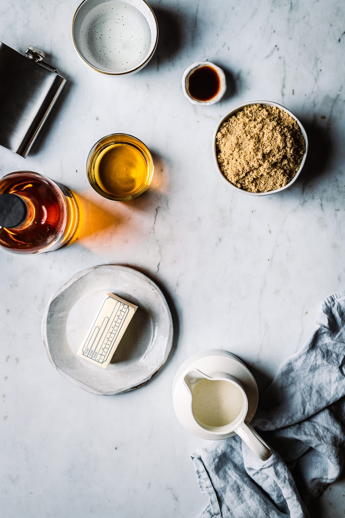 Ingredients for making caramels in containers on a grey marble surface. Light shines through a bottle of whiskey poured into a glass. Bowls of sugars and vanilla rest nearby along with a plate of butter and a pitcher of cream.