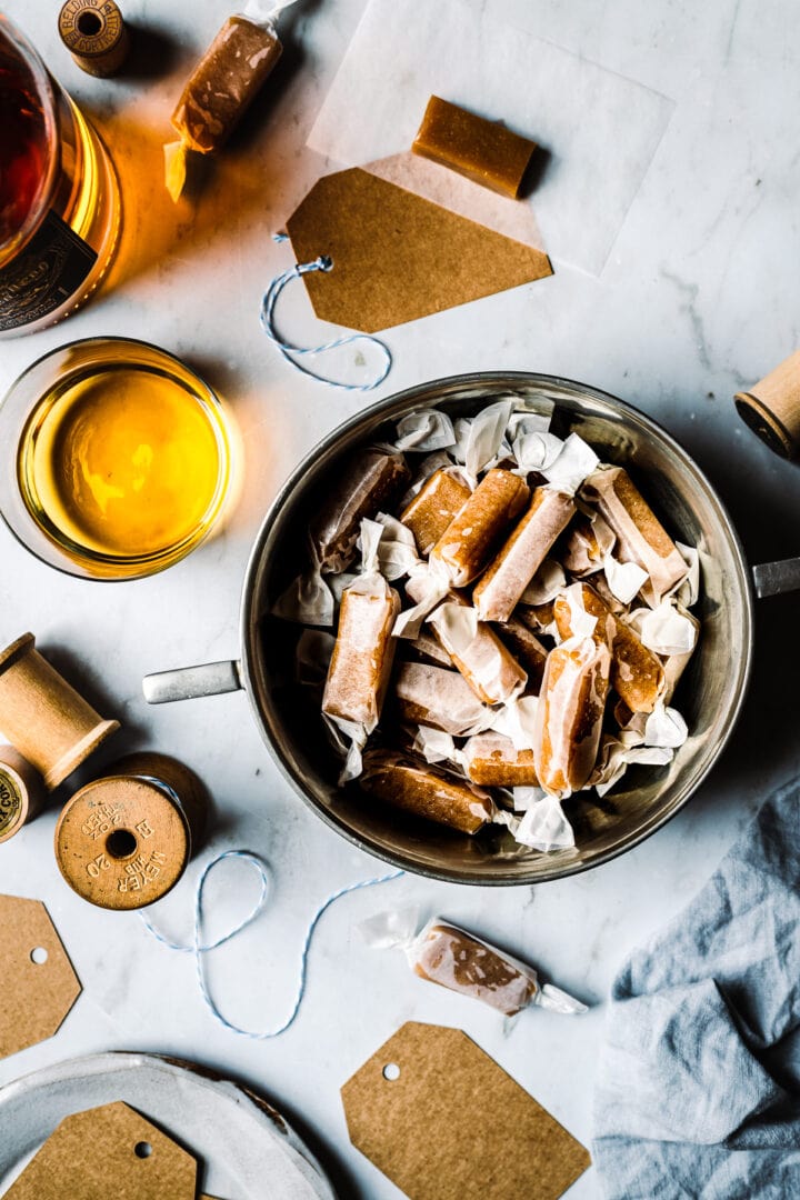 Pieces of candy wrapped in paper in a vintage silver bowl. The work surface is grey marble. There are paper candy wrappers, a vintage spool of twine, a blue linen napkin and gift tags nearby. Light shines through a whiskey bottle and glass.