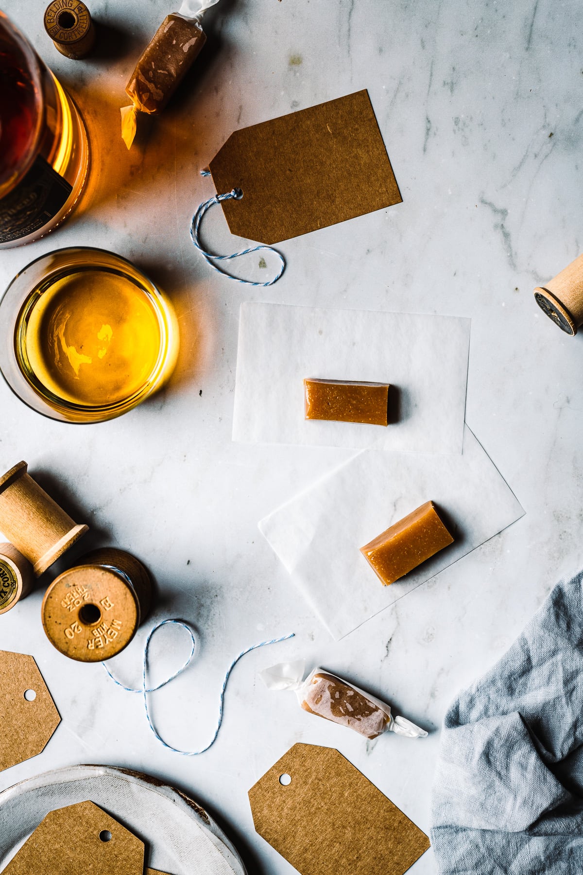 Whiskey salted caramels being wrapped on a grey marble surface. There is a vintage spool of twine, brown paper gift tags, and a blue linen napkin nearby.