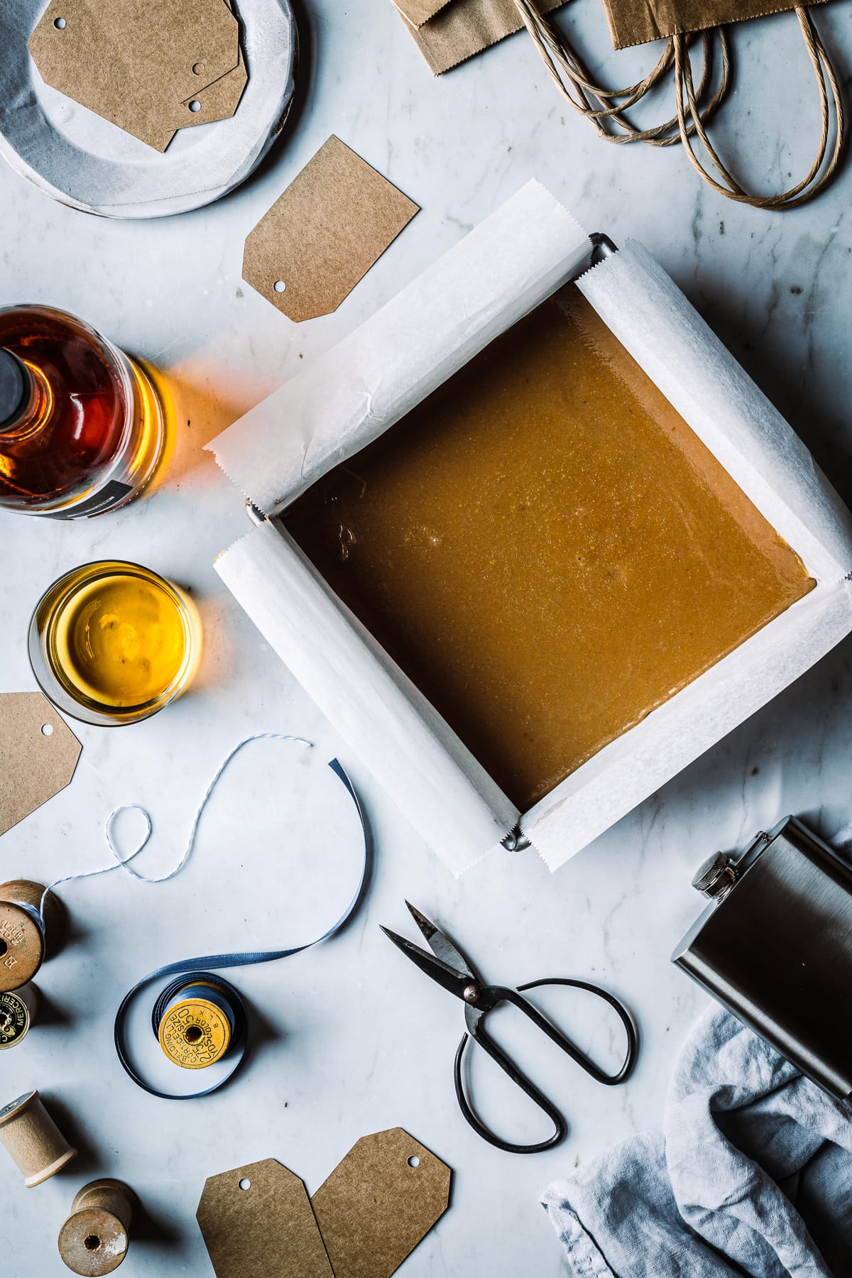 A pan of caramel on a cool marble surface with a bottle of whiskey and vintage wrapping supplies nearby