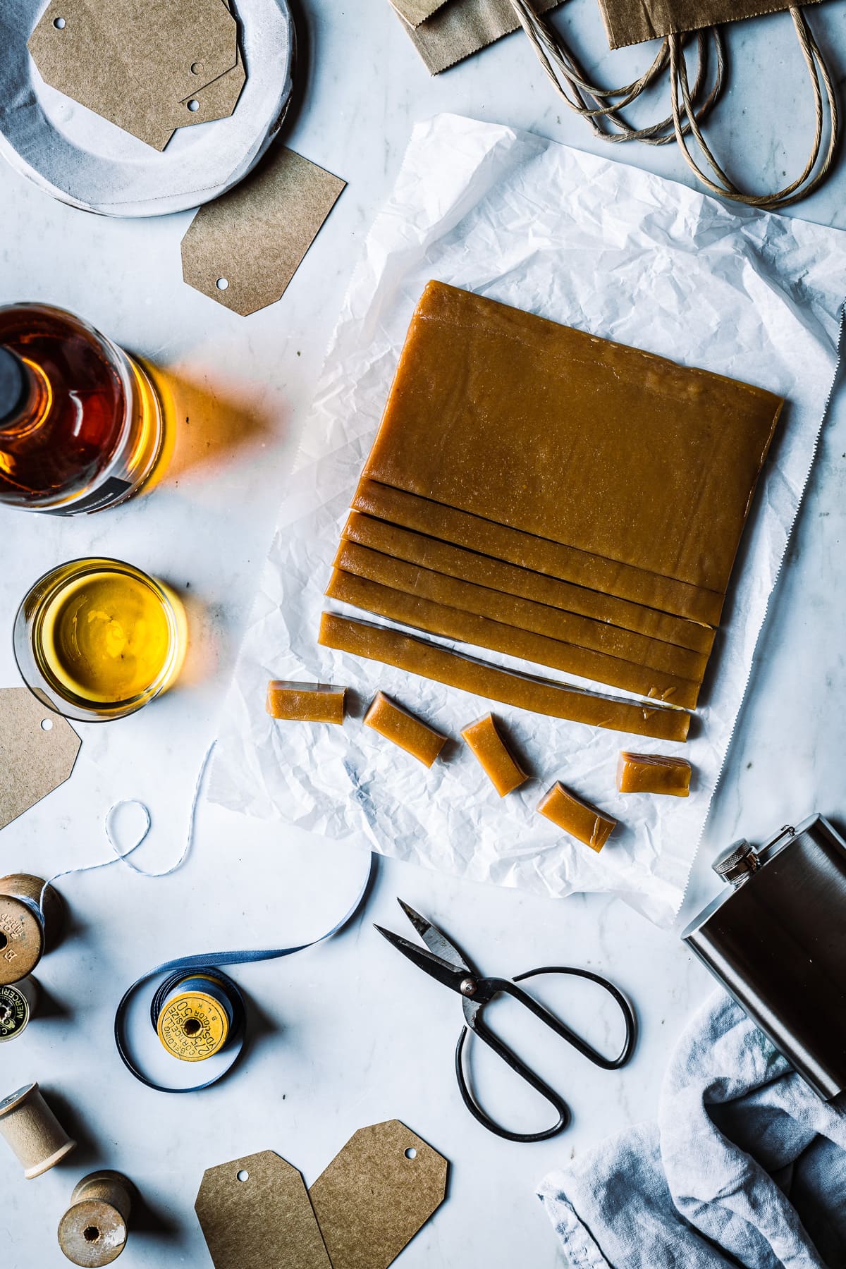 Caramels being cut into individual pieces on parchment paper on a marble surface. Surrounding the caramels are gift wrapping supplies, a bottle of whiskey, scissors, ribbons, gift tags and a flask.